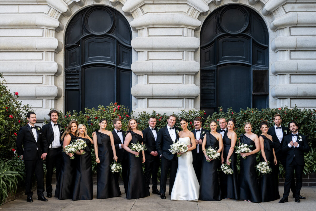 Group portrait of a wedding party at the Fairmont San Francisco. The groomsmen are all wearing black tuxes and the bridesmaids all in black. Photo by San Francisco Wedding Photographer Duy Ho