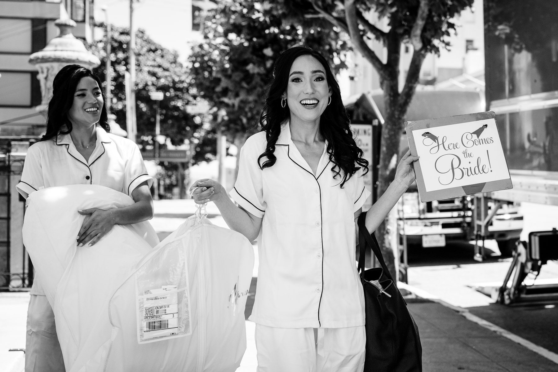 Black and white photo of bride and bridesmaid in matching pajamas carrying wedding dress on San Francisco street