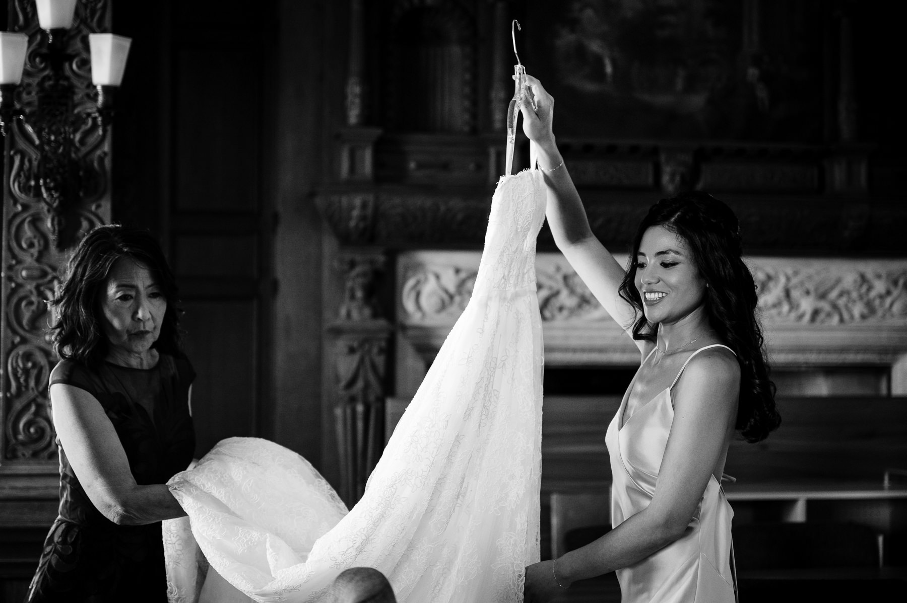 Black and white photo of bride holding up wedding gown in Flood Mansion library, mother watching, ornate woodwork behind