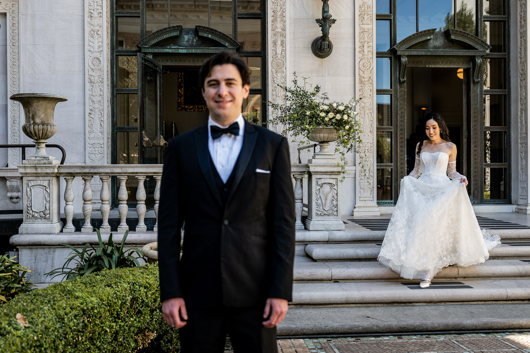 First look — groom in tuxedo waiting on Flood Mansion steps, bride approaching through arched doorway in background